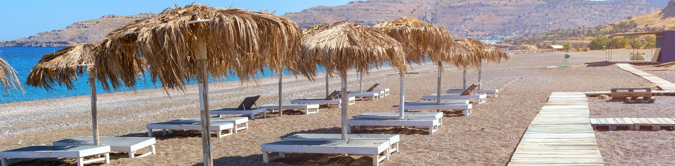 Mehrere Liegen mit Sonnenschirmen stehen auf einem Kiesstrand vor blauem Meer und klarem Himmel, im Hintergrund Felsen.