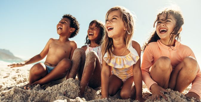 Gruppe fröhlicher Kinder sitzt im Sand am Strand und lacht während eines sonnigen Badetag-Urlaubs