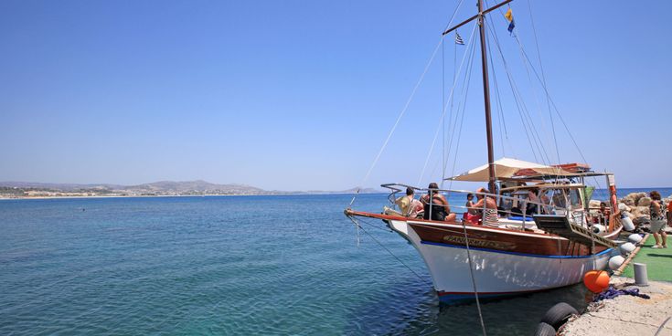 Mehrere Personen sitzen auf einem hölzernen Segelboot, das an einem Steg im Meer vor einer Küstenlandschaft festgemacht ist.