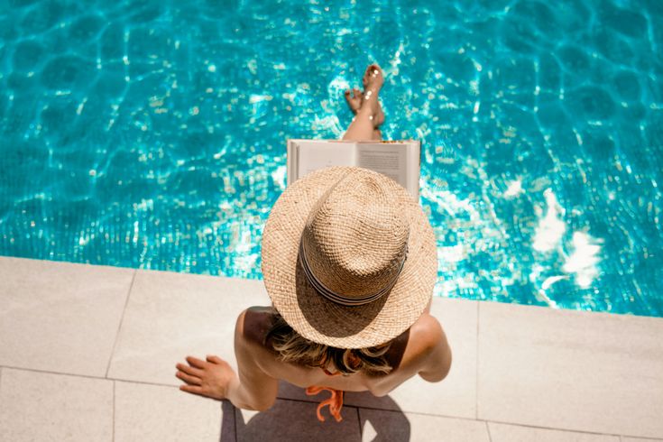 Person sitzt am Pool mit Strohhut und liest ein Buch, entspannte Sommeratmosphäre am Wasser