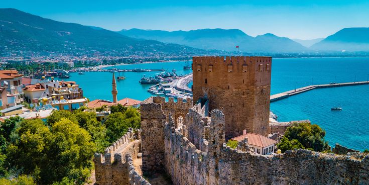 Historischer Roter Turm in Alanya mit Blick auf den Hafen und das tiefblaue Mittelmeer, umgeben von alten Festungsmauern und grüner Vegetation, im Hintergrund die Taurusgebirge