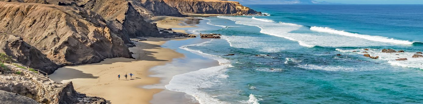 Strand Playa del Viejo Reyes auf Fuerteventura mit Sand, Felsen, drei Personen am Ufer und Wellen im Atlantik unter blauem Himmel mit Wolken.