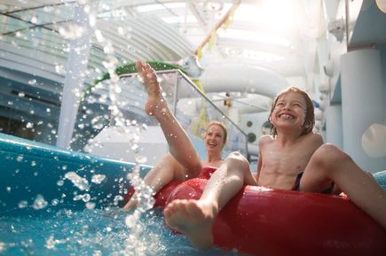 Personen rutschen auf einem großen Reifen durch den Lazy River der AIDAprima, während Wasserspritzer in der hellen, überdachten Wasserlandschaft aufsteigen