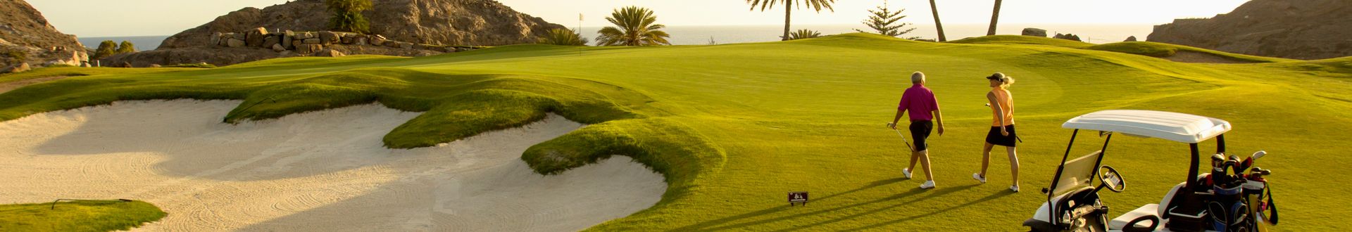 Personen gehen mit Golfschlägern über einen Golfplatz mit Sandbunkern und Golfcart vor einer Küstenlandschaft am Meer.