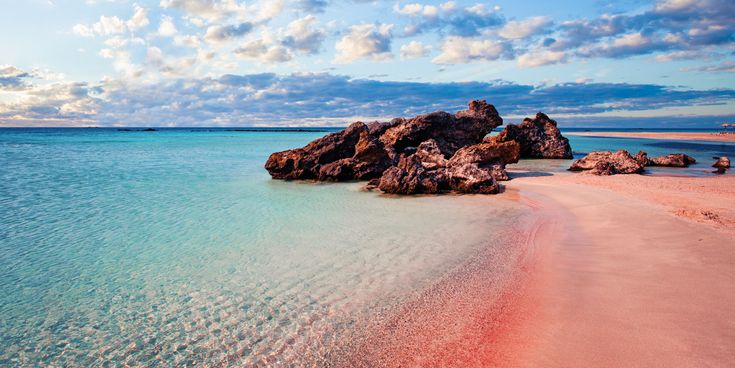 Klares türkisfarbenes Meer mit flachem Sandstrand und einer Gruppe brauner Felsen unter bewölktem Himmel.