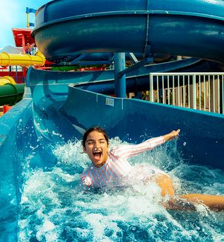 Mädchen in gestreiftem Shirt rutscht auf blauem Wasserrutschenkanal im Legoland Dubai Wasserpark, umgeben von bunten Wasserrutschen und Palmen.