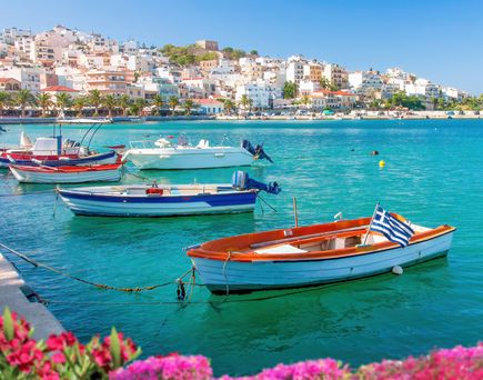 Mehrere kleine Boote mit griechischer Flagge liegen an einer Hafenmauer, im Hintergrund Häuser an einem Hang und blühende pinke Blumen im Vordergrund.