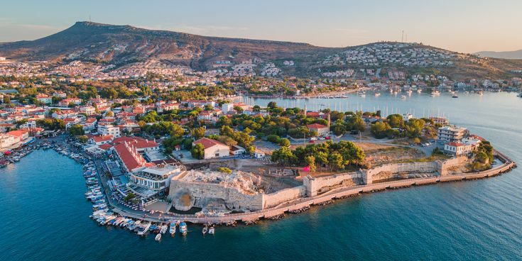 Luftaufnahme einer Küstenstadt bei Izmir in der Türkei mit Marina, alten Festungsmauern auf einer Landzunge und Hügeln im Hintergrund