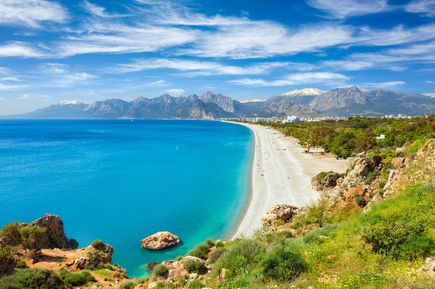 Weitläufiger Konyaalti-Strand in Antalya mit hellem Kies, klarem blauem Meer und dem Taurusgebirge unter einem sonnigen Himmel