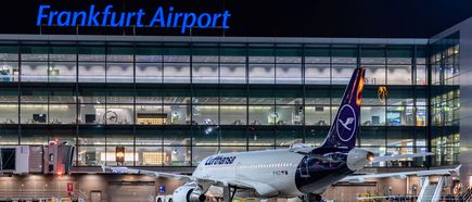 Lufthansa-Flugzeug auf dem Vorfeld des Flughafens Frankfurt bei Nacht vor der beleuchteten Glasfassade des Terminals mit gut sichtbarem Schriftzug „Frankfurt Airport“