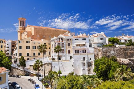 Die Stadt Mahón auf Menorca zeigt helle Häuser, Palmen und die historische Kirche Santa Maria unter blauem Himmel