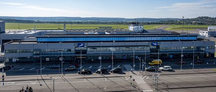 Frontansicht des Terminals am Flughafen Saarbrücken, modernes Flughafengebäude mit Vorfahrt, parkenden Fahrzeugen und Blick auf das Rollfeld bei Tageslicht
