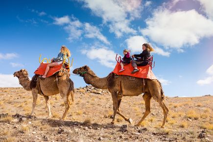 Zwei Frauen mit einem Kind reiten auf Kamelen durch eine trockene, hügelige Landschaft unter blauem Himmel mit leichten Wolken