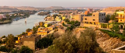 Blick auf Assuan am Nil mit traditionellen Segelbooten, modernen und alten Gebäuden sowie Wüstenhügeln im Hintergrund bei warmem Abendlicht