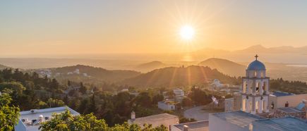 Landschaftsaufnahme von Kos mit Sonnenuntergang, Hügeln, Gebäuden und einer Kirche mit blauem Kuppeldach im Vordergrund