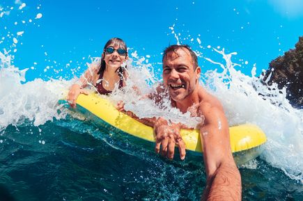 Mann und Kind mit Schwimmbrille auf gelber Luftmatratze im Meer mit spritzendem Wasser und Wellen