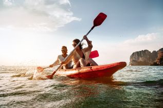Zwei Personen paddeln im roten Kajak auf dem Meer, mit Wasserspritzern und Felsen im Hintergrund