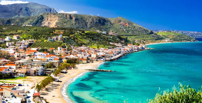 Blick auf Kalyves auf Kreta mit Strand, türkisfarbenem Meer, Häusern am Ufer und Bergen im Hintergrund.