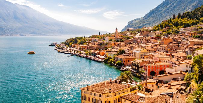 Blick auf eine belebte Uferstadt am Gardasee in Norditalien, umgeben von Bergen und klarem Wasser
