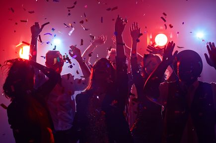 Menschen feiern in einem Club unter bunten Lichtern, werfen die Arme in die Luft, während Konfetti fällt