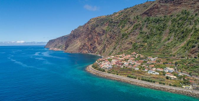 Küstenlinie mit steilem, bewaldetem Berghang und kleinem Dorf am Meer unter blauem Himmel