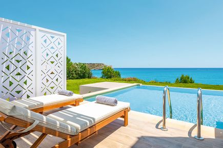 Zwei Holzliegen mit weißen Polstern und grauen Handtüchern neben einem Pool mit Meerblick und blauem Himmel.