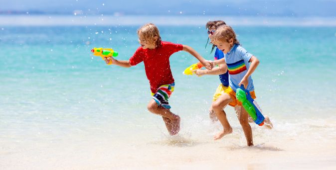 Kinder rennen lachend am Strand entlang und spritzen mit Wasserpistolen, während sie durch das flache Meerwasser spielen