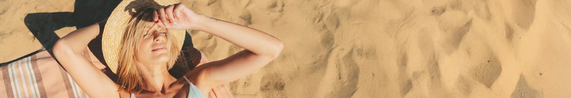 Frau liegt entspannt auf einem gestreiften Handtuch am Sandstrand und hält einen Strohhut über dem Gesicht, während sie die Sonne genießt