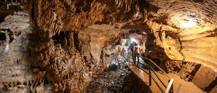 Beleuchteter Gang in einer Tropfsteinhöhle mit Felsformationen und einem schmalen Steg, auf dem zwei Personen gehen.