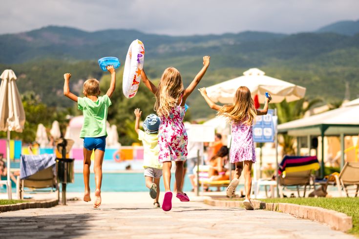 Mehrere Kinder laufen auf einer Hotelanlage in Richtung Poolbereich, umgeben von Liegen, Sonnenschirmen und einer Bergkulisse im Hintergrund
