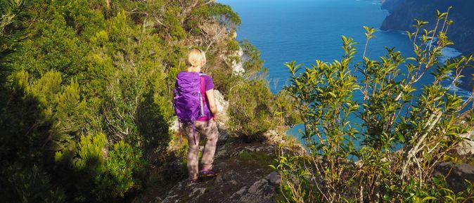 Person mit Rucksack steht auf einem Aussichtspunkt der Vereda do Larano auf Madeira und blickt über die bewaldete Steilküste und den Atlantik