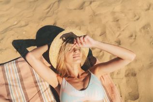 Person liegt entspannt auf einem gestreiften Handtuch im warmen Sand und hält einen Strohhut über dem Gesicht, während die Sonne die Strandfläche beleuchtet