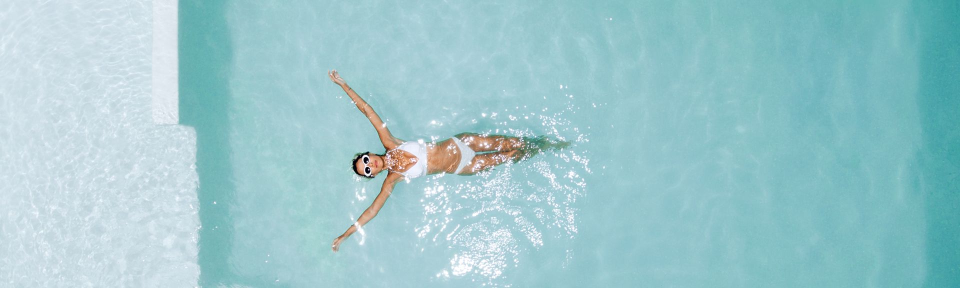 Frau in weißem Bikini und mit Sonnebrille liegt entspannt auf dem Rücken im hellblauen Pool.