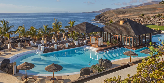 Blick auf einen Pool mit Sonnenschirmen und Liegen, umgeben von Palmen und einem Holzgebäude mit großen Fenstern am Meer.