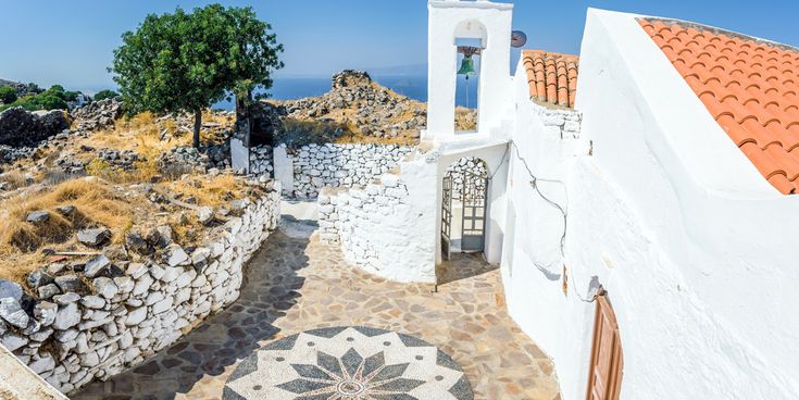 Weg mit weiß getünchten Mauern und Glockenturm neben weißem Gebäude mit rotem Ziegeldach auf felsigem Hügel mit Baum und Meer im Hintergrund