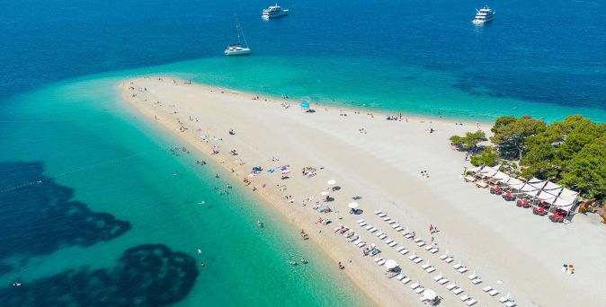 Luftaufnahme eines sandigen Strandes mit Liegestuhlreihen, Sonnenschirmen, Badegästen und mehreren Booten im türkisfarbenen Meer