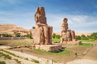 Zwei große, verwitterte Steinstatuen der Memnonkolosse in einer Wüstenlandschaft bei Luxor, Ägypten
