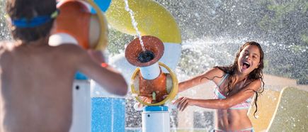 Zwei Kinder spielen lachend im Splash-Pad, Wasserfontänen und Spritzgeräte sorgen für Abkühlung in einer Ferienanlage