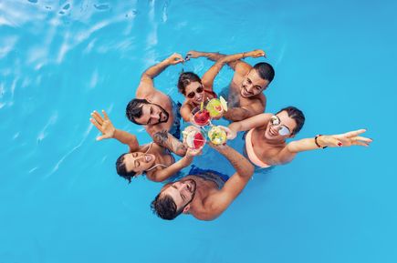 Gruppe junger Menschen im Pool stößt mit bunten Cocktails an und lacht in die Kamera