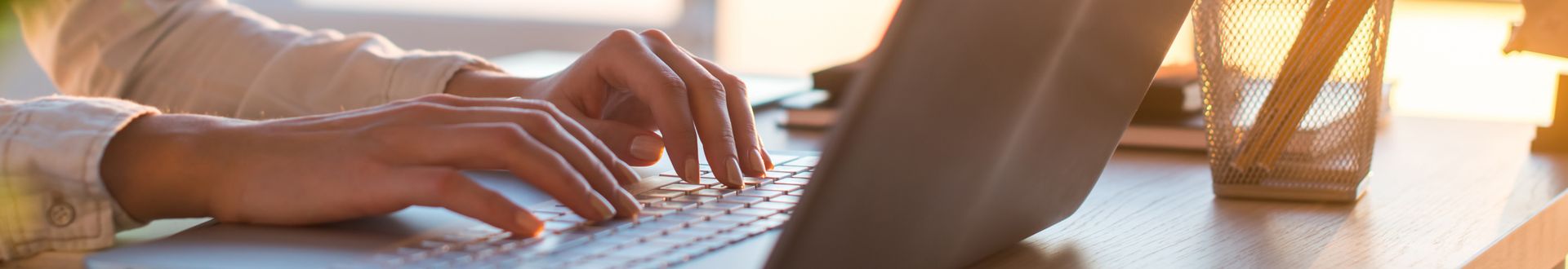 Nahaufnahme einer Person, die an einem Schreibtisch auf der Tastatur eines Laptops tippt, mit warmem Licht und Arbeitsutensilien im Hintergrund