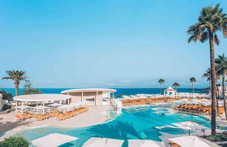 Großer Hotelpool mit mehreren weißen Sonnenschirmen und orangefarbenen Liegestühlen, Palmen und Meer im Hintergrund unter blauem Himmel.