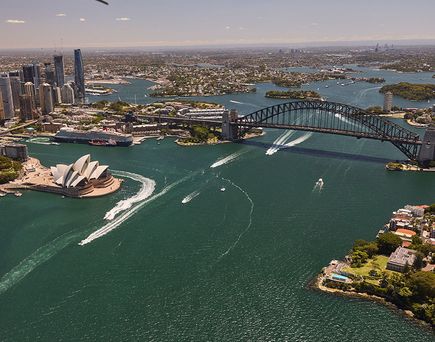 Luftaufnahme von Sydney mit Opernhaus, Hafenbrücke und mehreren Booten auf dem Wasser.