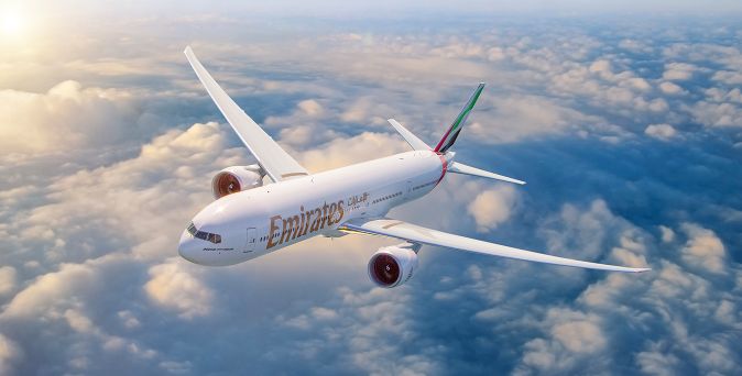 Passagierflugzeug der Fluggesellschaft Emirates fliegend über Wolken bei Sonnenuntergang, weiß mit rotem und grünem Logo am Heck.