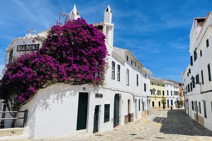 In Es Mercadal auf Menorca säumen weiße Häuser mit grünen Fensterläden eine schmale Straße, während violette Bougainvillea an einer Hauswand blüht