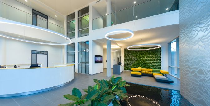 Heller Empfangsbereich mit rundem weißen Empfangstresen, großen runden Deckenleuchten, grüner Pflanzenwand und gelb-blauem Sitzbereich neben Wasserbecken