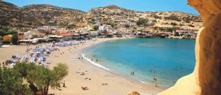 Blick von einer Felsöffnung auf einen Sandstrand mit mehreren Sonnenschirmen, Badegästen und einem Dorf am Hang.