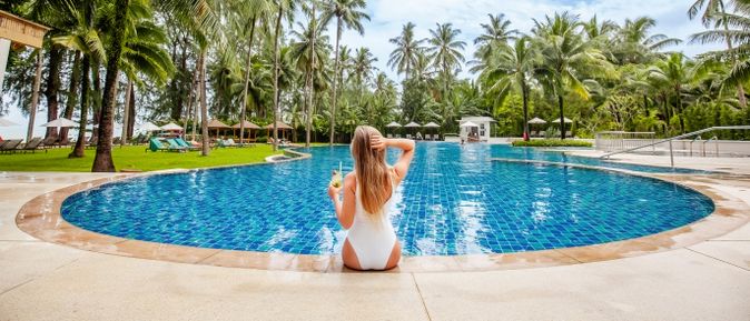 Frau in weißem Badeanzug sitzt am Rand eines großen Hotelpools und hält ein erfrischendes Getränk, umgeben von Palmen und Sonnenliegen in einer tropischen Ferienanlage