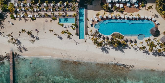 Luftaufnahme eines Resorts am Strand mit einem großen Pool, Liegen auf der Terrasse, Palmen und einem Sandstrand, der ins Meer übergeht.