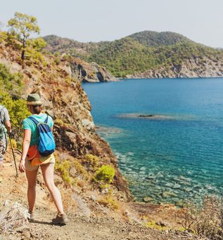 Ein Paar geht an der Küste wandern mit Rucksack und Wanderstöcken. Im Hintergrund sieht man das Meer und eine Bergkette