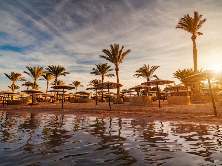 Strand mit Palmen und Sonnenschirmen in Hurghada bei Sonnenuntergang, der Sand und das ruhige Meer spiegeln warmes Licht wider